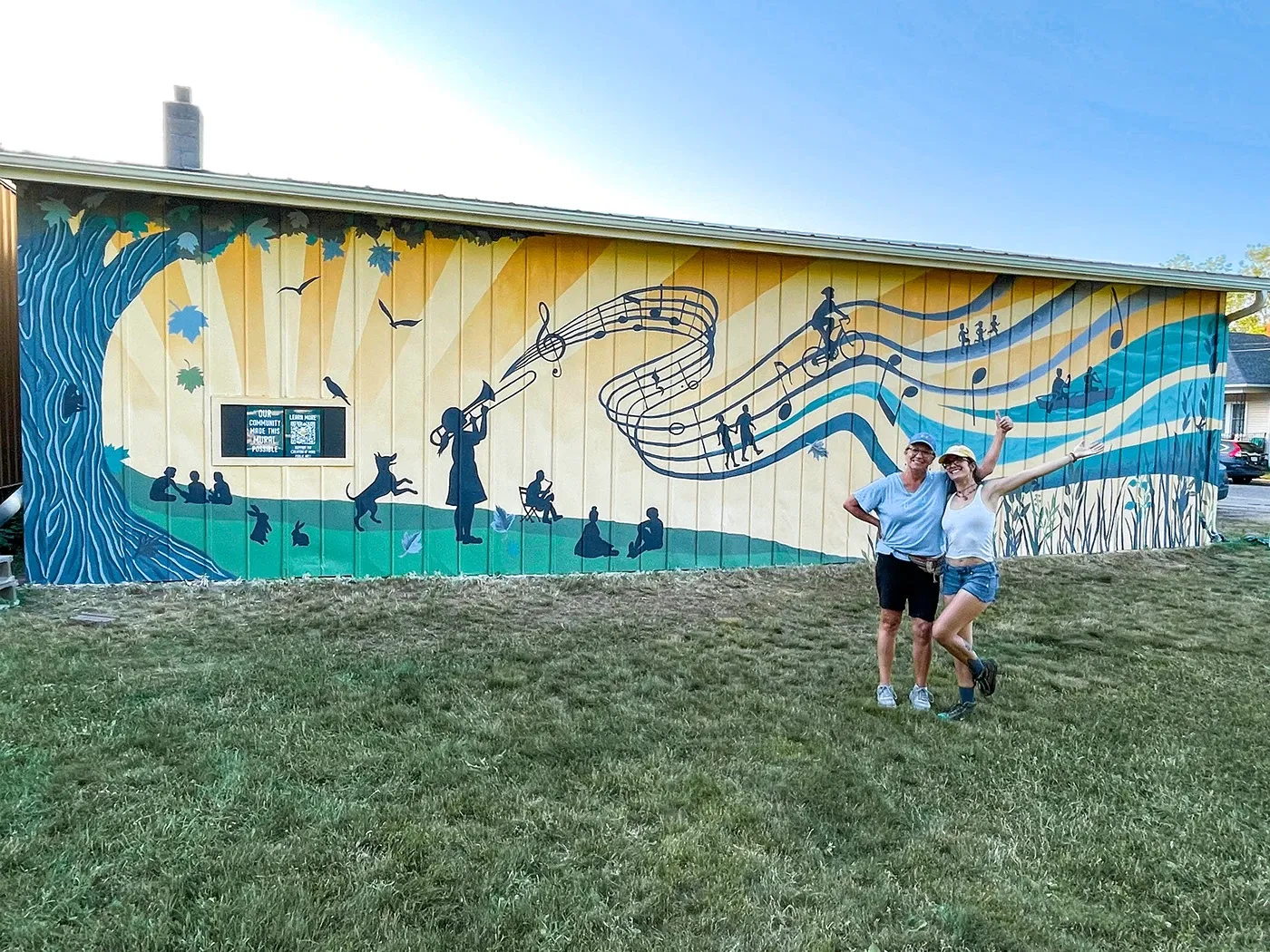 Revelry community mural on a building exterior: a flowing musical staff, silhouetted figures and animals, and a sunlit tree, with two artists standing in front.