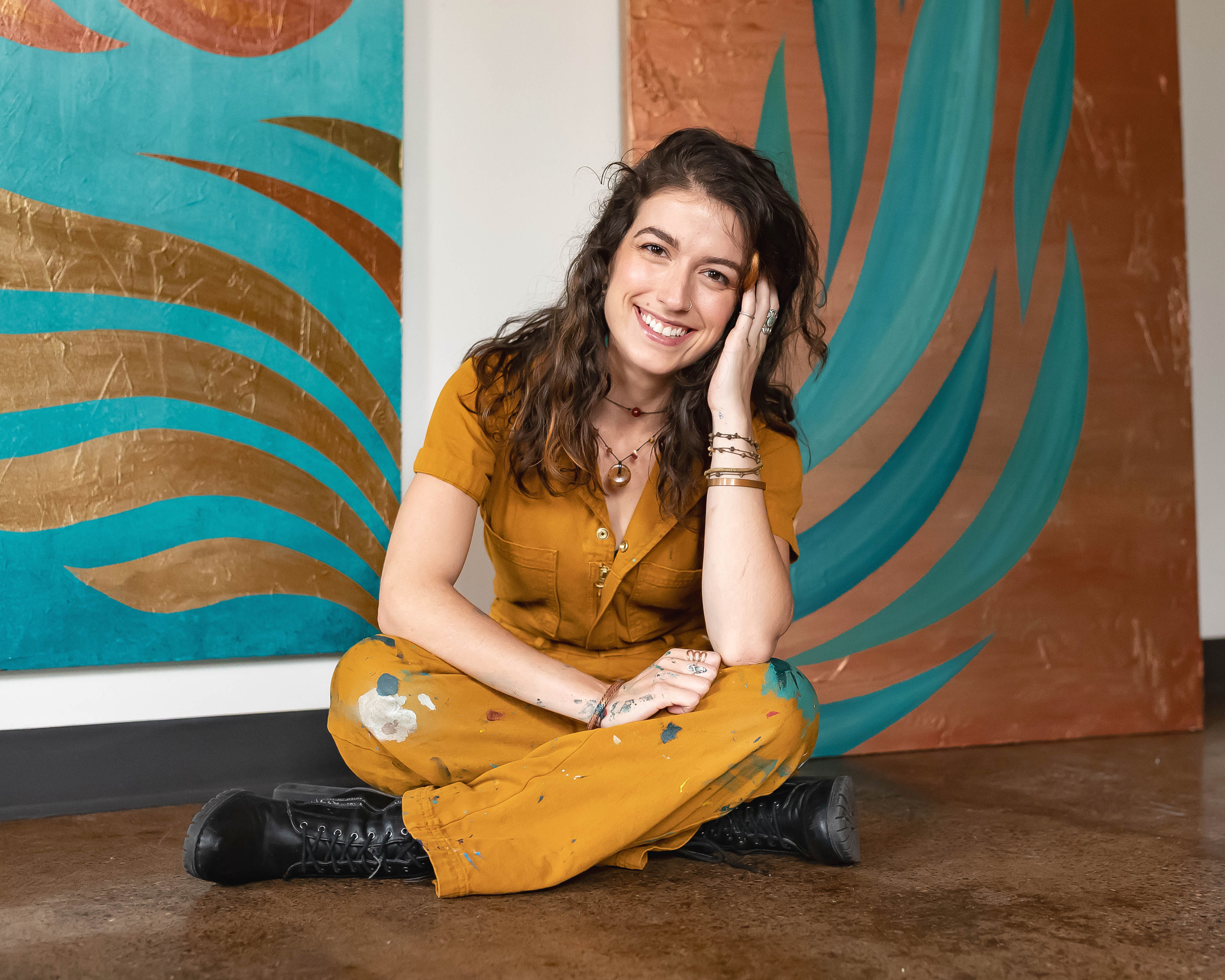 Sylvia Hecht seated in front of two of her mural panels, in a paint-flecked rust-colored jumpsuit.