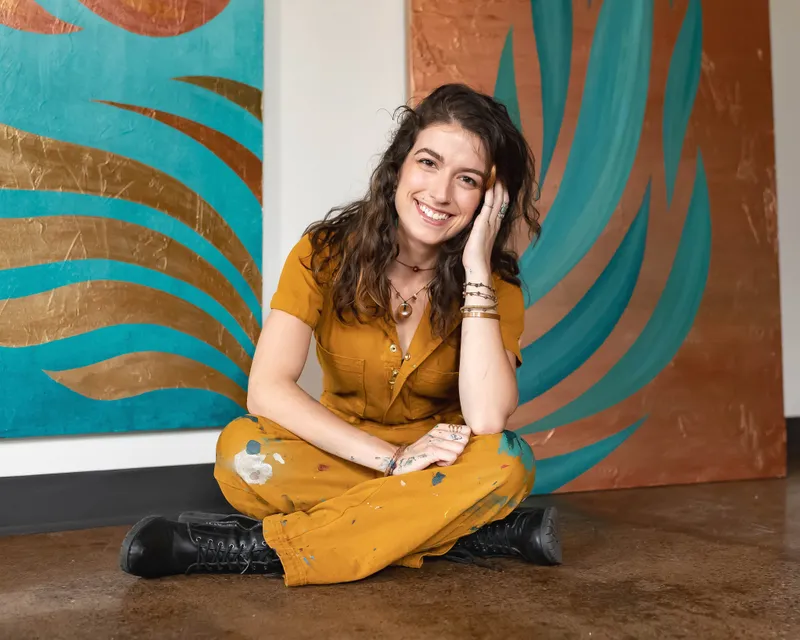 Sylvia Hecht seated in front of two of her mural panels, in a paint-flecked rust-colored jumpsuit.