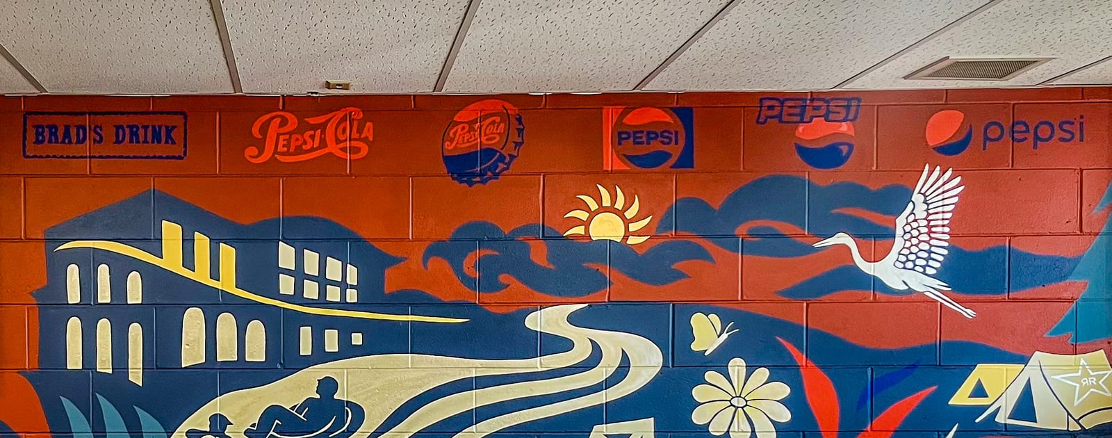 Eau Claire mural on a painted cinder block wall: a row of Pepsi wordmarks from the 1893 Brad’s Drink stamp through the modern lowercase logo, above an orange sky with a sun and clouds, a blue river winding past a bridge and downtown buildings, a heron in flight, a paddler on the water, and a daisy.