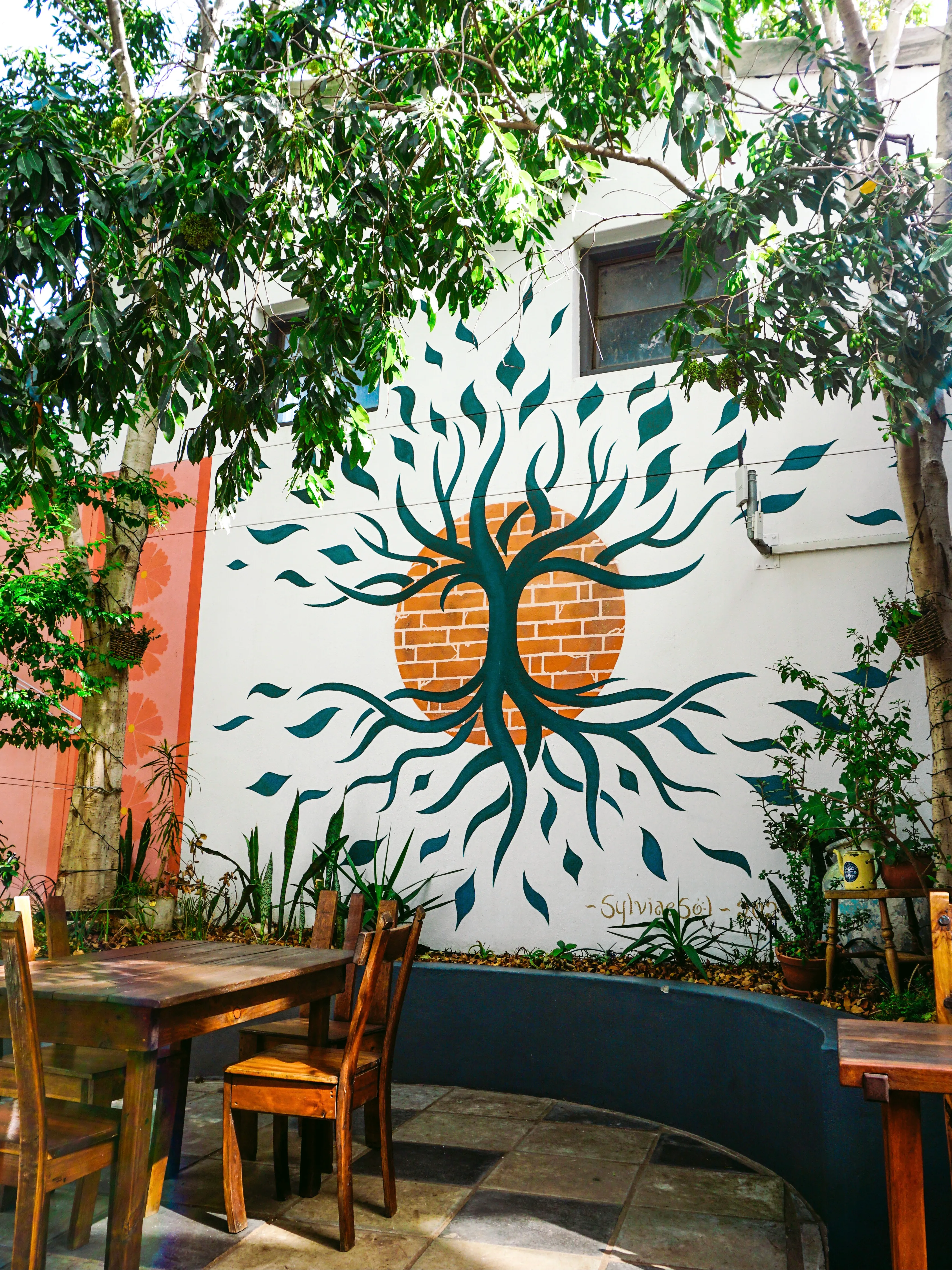 Tree-of-life mural on a white courtyard wall, branches and roots radiating from a brick-circle sun, with cafe tables below.