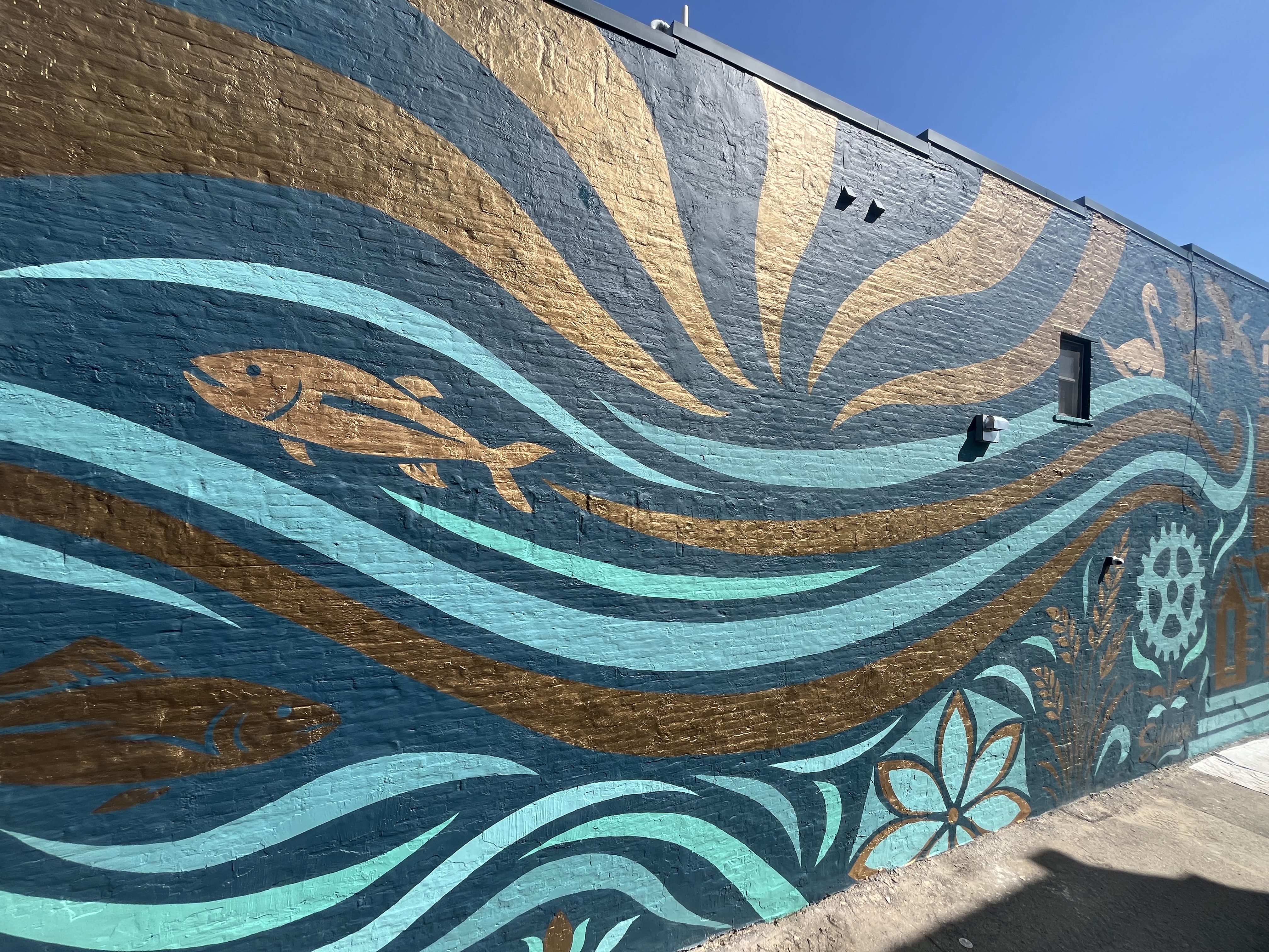 Exterior brick wall mural of sun rays over teal waves with copper-colored fish and seaweed motifs.