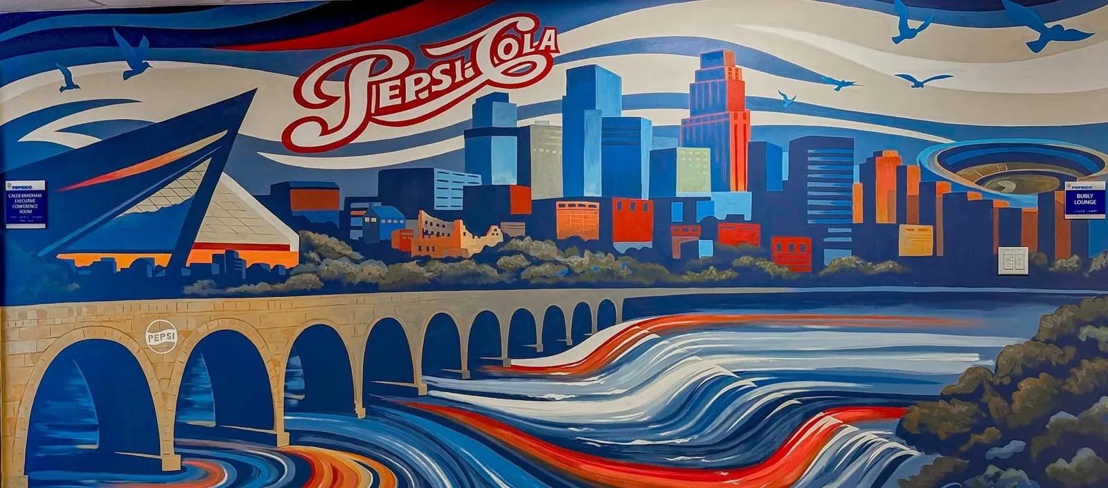 Mural of the Minneapolis skyline behind the Stone Arch Bridge, with US Bank Stadium at left, the river running in red, white and blue ribbons below, and retro Pepsi-Cola script across the sky.