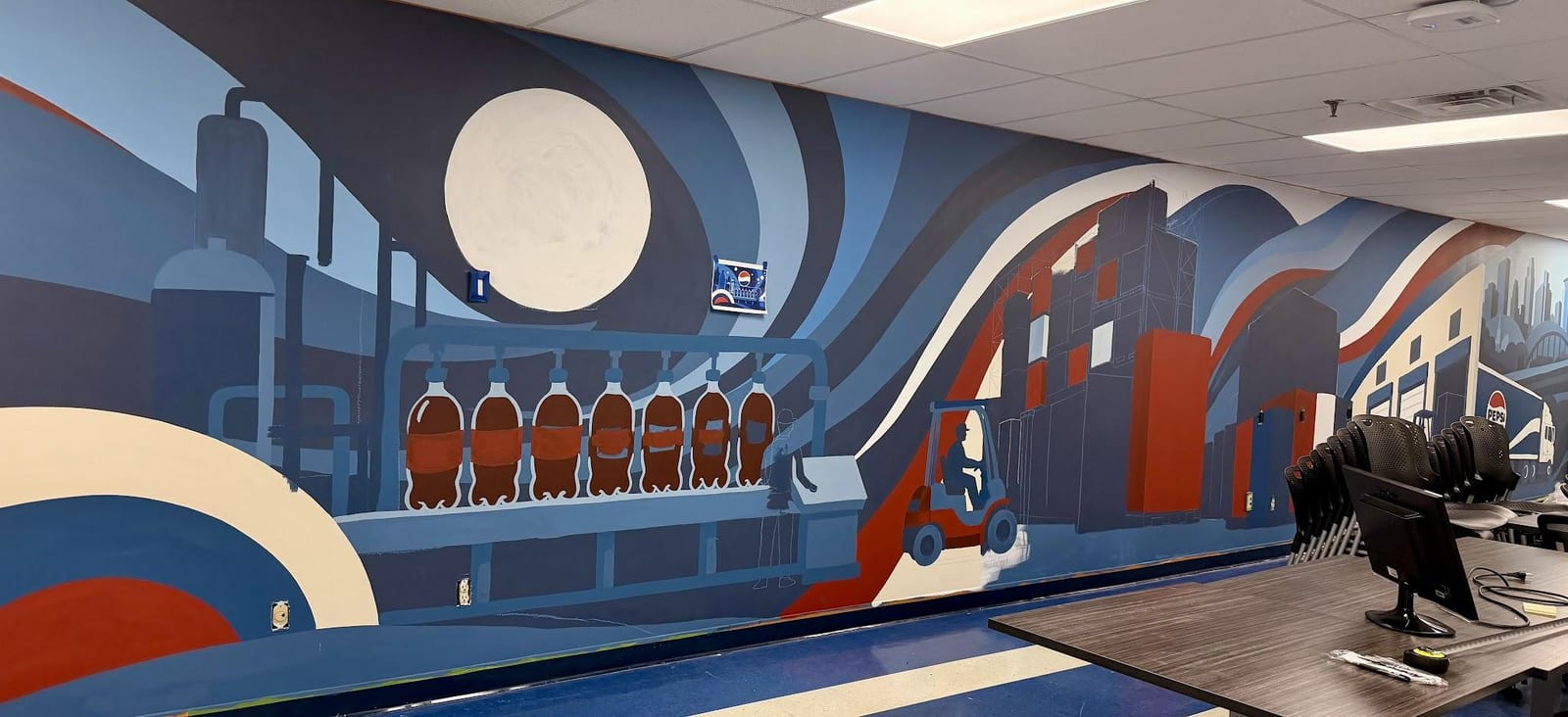 The production mural partway through installation in a training room, with the room’s tables, office chairs and a monitor still in place, drop cloths and tape along the baseboard, and one large circle still unpainted white.
