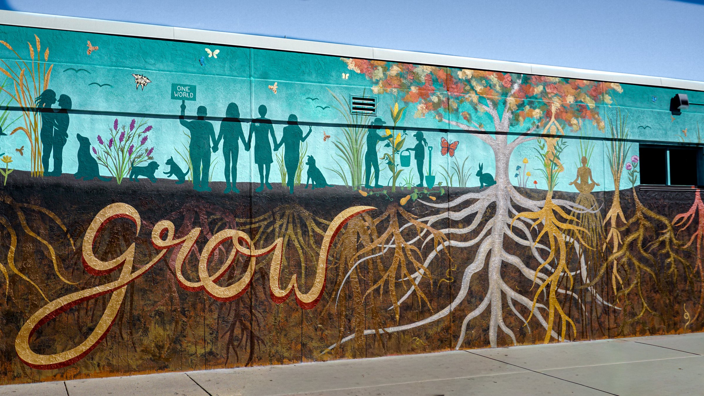 Full view of the Roots of Our Community mural: a gold 'grow' script, silhouetted neighbors holding hands, a tree, and white and gold root systems above and below ground.